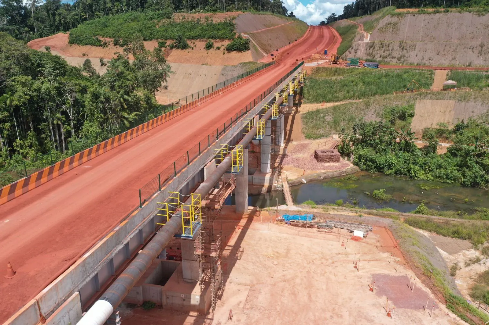 Ponte de concreto sobre curso d’água em estrada de terra vermelha, com floresta ao fundo