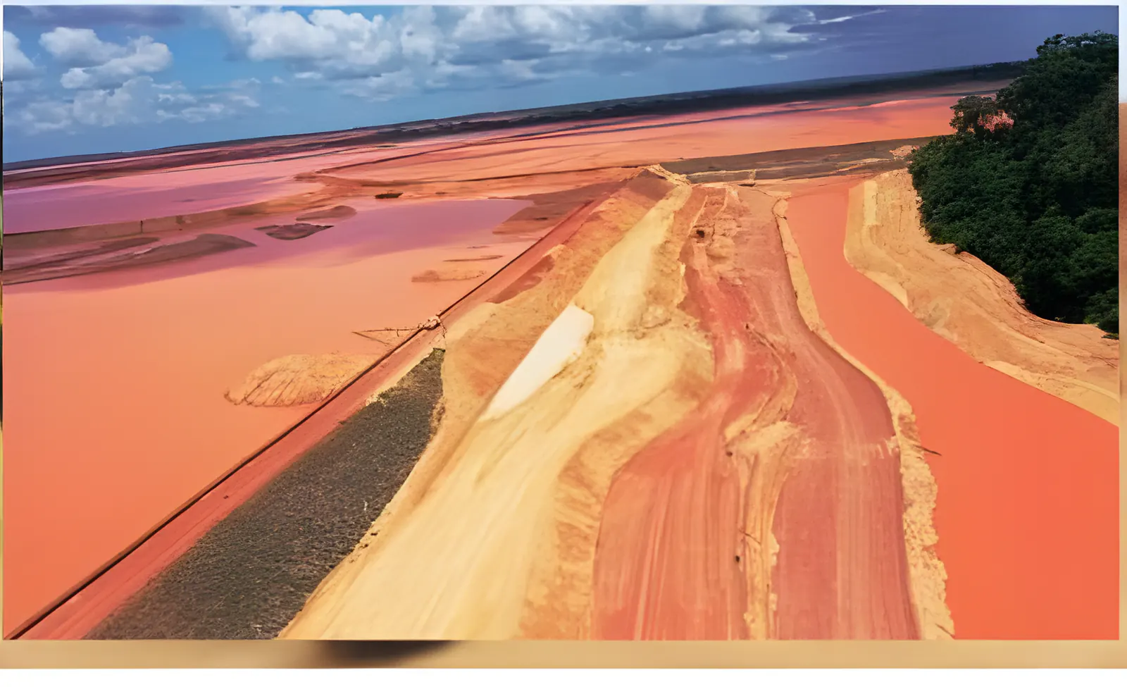 Vista aérea de berma em terra ao longo de reservatório de água avermelhada, com floresta à direita